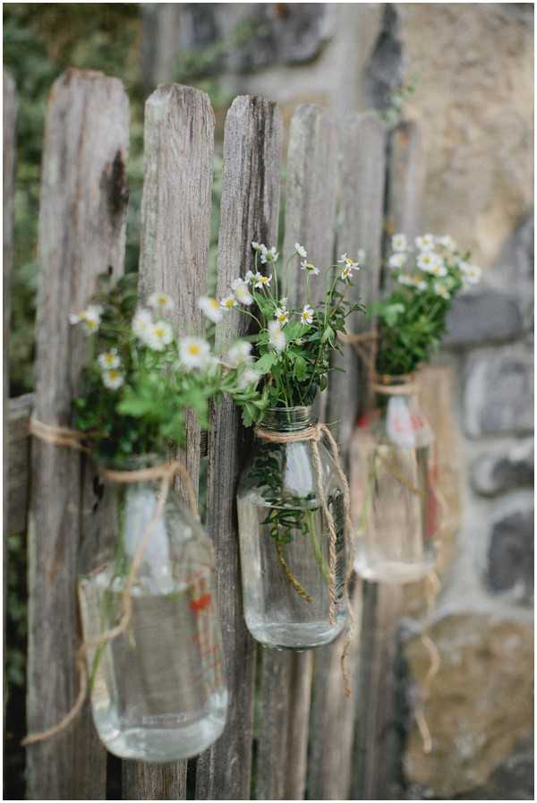 Close-up detail shot of rustic outdoor wedding decor featuring three clear glass bottles tied with natural twine and hung from a weathered wooden fence. Each bottle contains small white and yellow wildflower-style blooms with green foliage arranged in a loose, garden-picked style. The decor palette is natural and understated, consistent with a rustic or boho styling theme. The background shows a stone wall slightly out of focus.