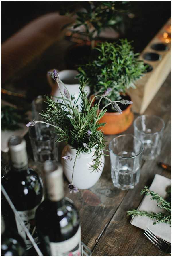 Close-up detail shot of a rustic wedding reception table styled with an herb-forward, garden-inspired theme. The centerpieces consist of fresh rosemary and lavender sprigs arranged in a small white ceramic pitcher and a terracotta pot, with additional herb cuttings scattered on the table and tucked beside napkins. Multiple bottles of red wine are grouped at the left side of the frame, alongside simple faceted drinking glasses and silver forks placed on linen napkins. The table surface is raw, unfinished wood, and a wooden tealight holder with warm candlelight is visible in the background, contributing to the overall rustic, informal aesthetic.