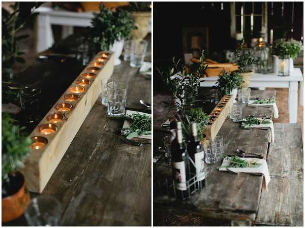 A diptych of reception table detail shots in a rustic indoor barn or farmhouse setting. The left image is a close-up of a raw wooden beam candle holder running the length of a reclaimed wood trestle table, with a row of lit tealight candles set into drilled holes and loose greenery scattered alongside glassware. The right image shows a wider view of the same table, revealing place settings with linen napkins, dark cutlery, glass tumblers, wine bottles in a wire caddy, and abundant fresh greenery including olive branches and herbs used as both centerpieces and plate garnish. The overall decor palette is dark and natural — raw wood, matte black accents, and deep green botanicals — consistent with a rustic or foraged styling theme with no flowers, relying entirely on candle warmth and greenery for decoration.