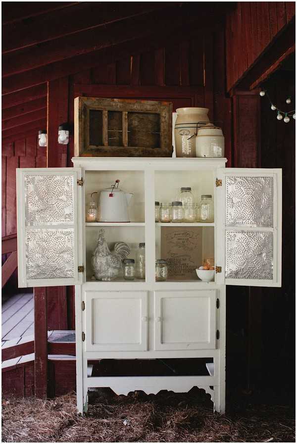 A close-up detail shot of a vintage white pie safe cabinet with punched tin door panels, styled as part of rustic wedding decor. The cabinet is open and displays mason jars, a white enamel percolator, ceramic rooster figurines, a small white bowl, and a printed sign, with a stoneware crock and a weathered wooden window frame displayed on top. The setting is inside or against a red barn structure, with string globe lights visible in the background. The overall styling theme is rustic farmhouse, using repurposed antique furniture and vintage kitchenware as decorative elements.