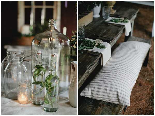 Close-up detail shots of rustic outdoor wedding reception decor, presented as a diptych. The left image shows a table styled with assorted clear glass vessels — a large demijohn, swing-top bottles with fresh green herb or vine stems, mason jars, and a small lit votive candle on a white linen tablecloth, photographed in warm ambient light against a dark background. The right image shows a weathered dark wood farm-style bench topped with a long cushion upholstered in natural linen ticking stripe fabric in cream and grey, with a rustic wooden table behind it holding greenery bundles, glassware, and a white linen runner. The overall styling is rustic-organic with a green and natural palette, using raw materials and handmade-feeling details throughout.