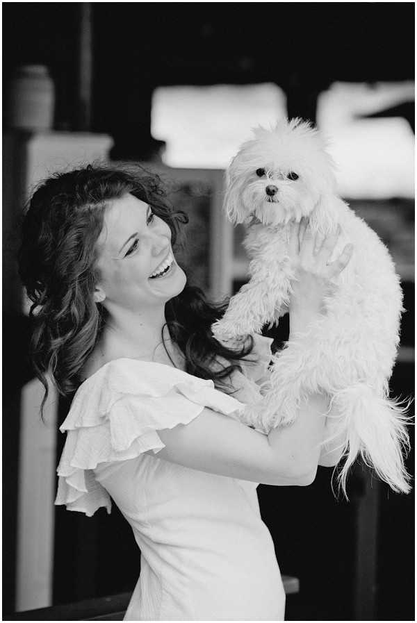 A black-and-white portrait of a bride holding a small white fluffy dog, likely a Maltese, up at eye level and laughing. The bride wears a white off-the-shoulder dress with tiered ruffle detailing and has long wavy dark hair worn loosely down. The image has bright highlights on the bride and dog contrasted against a dark, blurred indoor background. Close-up portrait composition with a shallow depth of field.