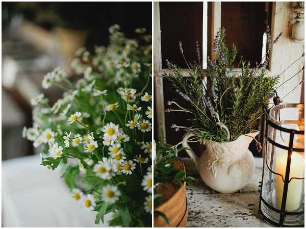 A split close-up detail shot of rustic wedding decor. The left panel shows a loose arrangement of small white chamomile-style daisy flowers with yellow centers and green foliage, placed on a white linen surface. The right panel features a white ceramic pitcher filled with rosemary sprigs, lavender stems, and other greenery, styled alongside a black metal-framed glass lantern holding a lit pillar candle. The overall decor palette is natural and herb-garden inspired, with a rustic, countryside aesthetic.