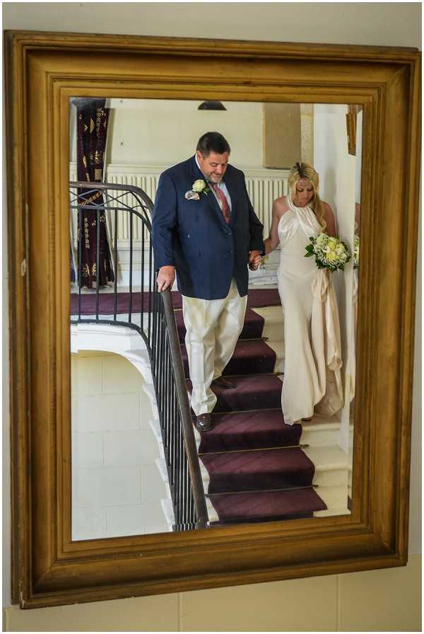 A creative portrait shot captured through the reflection of a large gold-framed mirror mounted on a wall, showing a bride and her escort descending a grand interior staircase with a deep burgundy carpet runner and wrought-iron railing. The man, likely the father of the bride, wears a navy double-breasted blazer, cream trousers, and a white boutonnière, and is holding the bride's hand as he leads her down. The bride wears a sleek ivory halter-neck gown with a minimalist silhouette and carries a round bouquet of ivory and pale yellow flowers with green foliage. The interior setting appears to be a chateau or manor house, with classic white stone walls and ornate architectural details visible in the background. The composition uses the gilded mirror frame as a creative framing device, adding depth to the image.