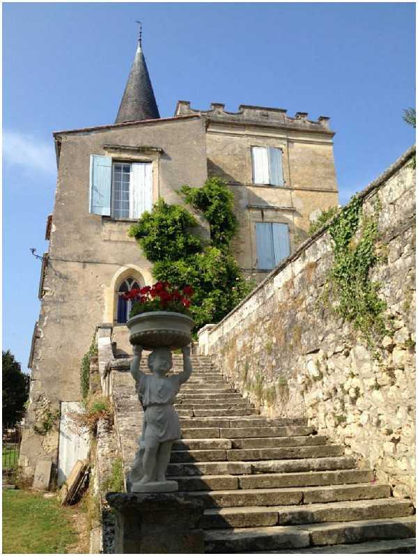 A wide shot of a historic French chateau or manor house exterior, featuring a stone staircase leading up to the main building. In the foreground, a decorative stone statue of a figure holds a large planter bowl filled with red geraniums. The building has pale blue shuttered windows, a pointed slate turret, and stone crenellations along the roofline, with climbing green ivy and shrubs framing the facade. No people or wedding-specific activity are visible in this image, which appears to be a venue exterior shot taken in natural daylight. Potential venue feature image.