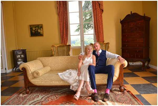 A bride and groom are seated together on an antique cream damask sofa inside a chateau salon, posing for a couple portrait. The bride wears a short white satin slip-style dress with thin straps and blush heels, while the groom wears a navy blue suit with a blue tie and pink socks. The room features yellow painted walls, terracotta and cream checkered tile flooring, a Persian rug, coral-orange floor-length curtains framing tall French doors, a dark wood chest of drawers, and a small ornate stove in the corner. A framed painting hangs on the wall above the sofa. The shot is a wide portrait-style image taken at mid-distance. Potential venue feature image.