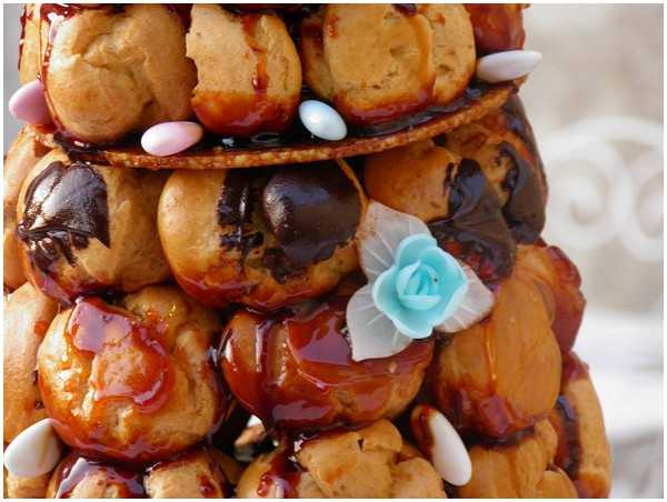 Close-up detail shot of a traditional French croquembouche wedding cake, showing cream puffs stacked in a tiered tower formation and held together with caramel and chocolate drizzle. The profiteroles are decorated with pastel Jordan almonds (dragées) in pink, blue, and white, and a small light blue sugar rose with white leaves is attached to one of the middle tiers. The background is softly blurred, suggesting an outdoor setting with white furniture visible.