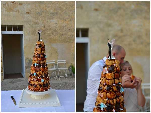 A side-by-side image showing a traditional French croquembouche wedding cake and the couple eating from it. The croquembouche is a tall cone-shaped tower of caramel-glazed profiteroles decorated with chocolate-dipped choux puffs and small pastel blue and pink sugar decorations, topped with a figurine bride-and-groom cake topper, displayed on a white square cake stand on a white linen-covered table with a cake knife beside it. The right panel shows a close-up portrait of the groom, who is bald and wearing a white shirt, leaning over the bride as they both eat a profiterole directly from the tower. The setting is an outdoor courtyard or terrace against a stone wall, consistent with a French chateau or farmhouse venue, suggesting a relaxed reception atmosphere.