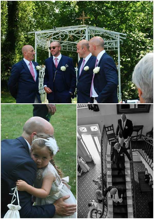 A three-image collage from a wedding day. Top image: four men in matching navy suits with pink ties and white rose boutonnieres stand and converse in front of a white ornate wrought-iron arch topped with a cross, set in an outdoor garden ceremony space — a medium wide shot. Bottom left: a man in a navy suit crouches to hug a young flower girl wearing a white dress and a white feathered headband, who is holding a small white drawstring bag — a close portrait shot taken outdoors on grass. Bottom right: a black-and-white aerial shot looking down a grand interior staircase with a wrought-iron banister, showing several formally dressed guests and wedding party members descending the stairs inside what appears to be a chateau or manor house — high contrast tones with bright natural light from a skylight window visible. Potential venue feature image.
