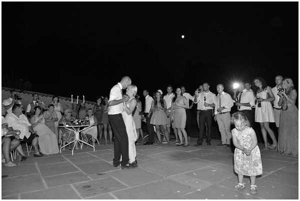 Black-and-white image of a first dance taking place on an outdoor stone terrace at night, with the couple embracing and kissing at the center of a circle formed by approximately 25-30 guests watching around them. The groom wears dark trousers and a light shirt, while the bride appears to be in a short white dress. A young girl in a patterned dress stands in the foreground slightly apart from the crowd. Guests are dressed in summer attire including short dresses and shirtsleeves, suggesting a warm evening event. A small cocktail table with candles and what appears to be a floral centerpiece is visible to the left, and several guests are photographing the moment with cameras and phones. The shot is a wide-angle, slightly elevated view capturing the full scene with high contrast between the dark sky and the lit figures below.