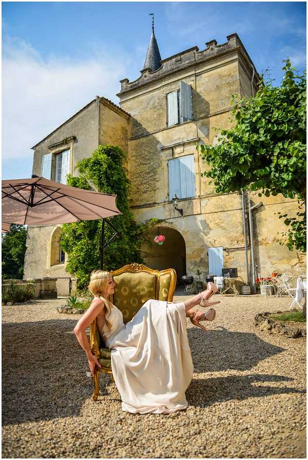 A bride in a white wedding dress and nude heeled sandals sits in an ornate gold-framed armchair upholstered in olive green damask fabric, posed in a playful position with her legs kicked up in the air. The portrait is shot outdoors in the gravel courtyard of a French chateau, a multi-story limestone building with pale blue shutters, a pointed turret, and climbing vines across the facade. The setting has a classic French country aesthetic, with a large taupe market umbrella, wrought iron garden chairs, and a table visible in the background. The wide shot captures both the bride and the full chateau exterior in bright midday sunlight. Potential venue feature image.