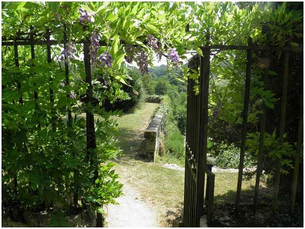 An outdoor garden setting photographed through an open wrought-iron gate covered with flowering wisteria in soft purple blooms and dense green foliage. The wide shot looks through the gateway onto a garden path leading to a low stone balustrade wall, with rolling countryside visible in the background. No people are present in the frame. The image primarily showcases the venue's garden architecture and grounds. Potential venue feature image.
