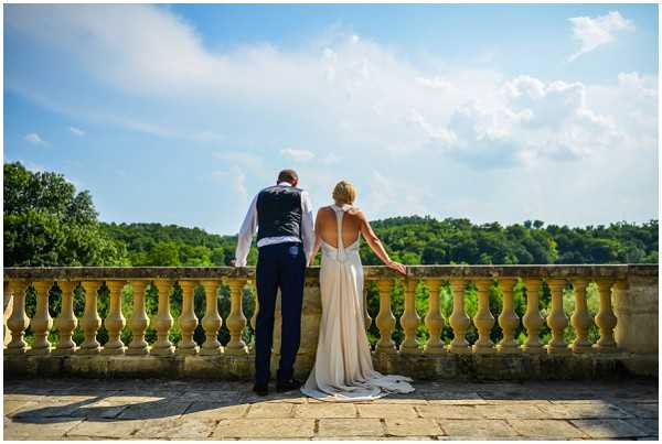 A couple stands side by side with their backs to the camera on a stone terrace with a classical balustrade, looking out over a wooded landscape. The groom wears navy trousers, a white dress shirt, and a navy patterned waistcoat, while the bride wears a champagne/ivory fitted gown with a low open back and halter neckline, with a short train resting on the stone floor. The shot is taken from behind in a wide portrait composition, emphasizing the open-back dress detail and the terrace architecture. Potential venue feature image.