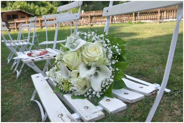 Close-up detail shot of a bridal bouquet resting on a white wooden folding chair at an outdoor wedding ceremony. The bouquet is composed of white roses, white calla lilies, baby's breath, green hypericum berries, and broad green leaves. Rows of white folding chairs are arranged on a grass lawn in the background, with ceremony programs visible on one of the chairs. A wooden structure, likely a chalet or pavilion, is visible in the soft-focus background, suggesting a garden or parkside setting. The overall decor palette is white and green with a classic, understated style.