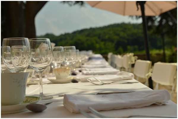 A close-up, ground-level shot of a long outdoor wedding reception table set for a large number of guests, extending into the background. The table is dressed with a white linen tablecloth and features place settings including white folded cloth napkins, silver cutlery, clear crystal wine glasses in multiple sizes, and small white ceramic bowls. White chairs are visible along the side of the table. A large white parasol is partially visible on the right side, and the setting appears to be a terrace or garden with a wooded hillside visible in the background.