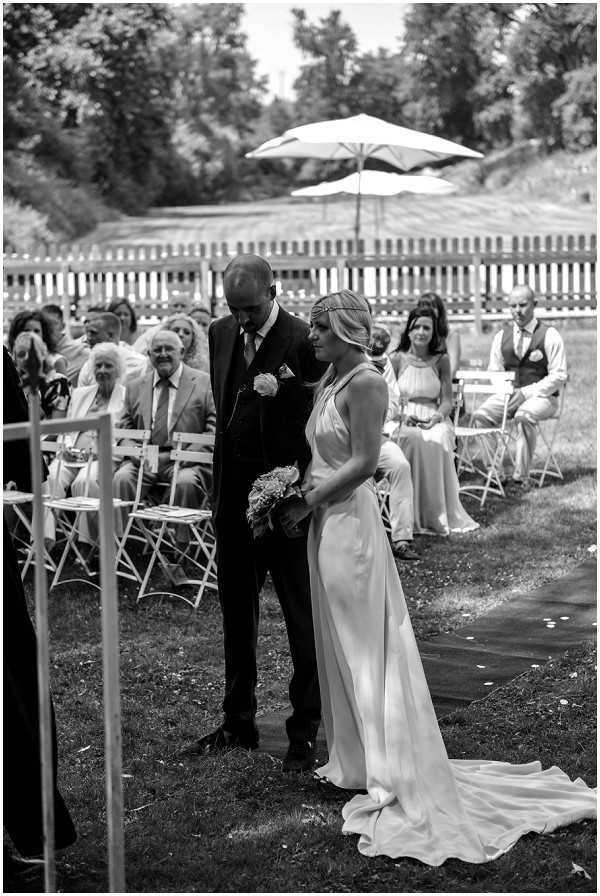 This black-and-white image captures an outdoor wedding ceremony taking place on a lawn, with the couple standing at the altar end of the aisle. The bride wears a sleek, fluid gown with a halter neckline, a long train, and a delicate headpiece or crown, and holds a small bouquet. The groom is dressed in a dark suit with a boutonniere. Both are looking downward, appearing to share a quiet moment. Behind them, approximately 20–25 guests are seated on white folding chairs arranged in rows on either side of a dark aisle runner. A bridesmaid in a light-colored halter dress is visible in the background, along with a groomsman in a waistcoat. A white picnic-style fence and a large patio umbrella are visible in the background. The image is a medium-wide shot taken from the officiant's side, capturing the full scene with strong contrast between the bright daylight and the dark tones of the groom's suit.