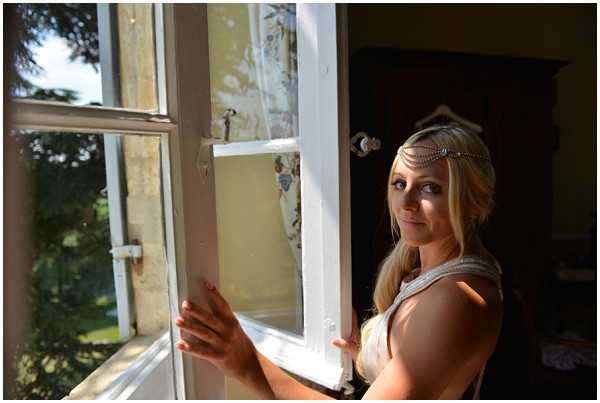 A getting-ready portrait of a bride standing at an open white-framed sash window inside what appears to be a chateau or manor house room, turning to look back toward the camera. She has long blonde hair worn loosely and is accessorized with a delicate silver chain headpiece across her forehead in a boho style. She is wearing a light grey or pale wrap top or getting-ready robe. The room behind her shows aged, peeling wallpaper with a floral pattern and a dark wood armoire, suggesting a vintage or rustic interior. Natural sunlight streams through the window, creating strong contrast between the bright window side and the darker interior. Close-up portrait composition with the window frame as a leading element.