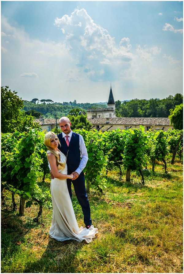 A couple portrait shot outdoors in a vineyard, with the bride and groom standing among rows of grapevines. The bride wears a sleeveless ivory/champagne draped gown with a small train and has long blonde hair worn loose; the groom wears a navy waistcoat over a white dress shirt with a pink tie and dark trousers. They are posed close together, both facing the camera, in a mid-length portrait composition. In the background, a French chateau or manor house with a pointed turret is visible among the vines, along with surrounding trees. The overall styling is classic and understated.