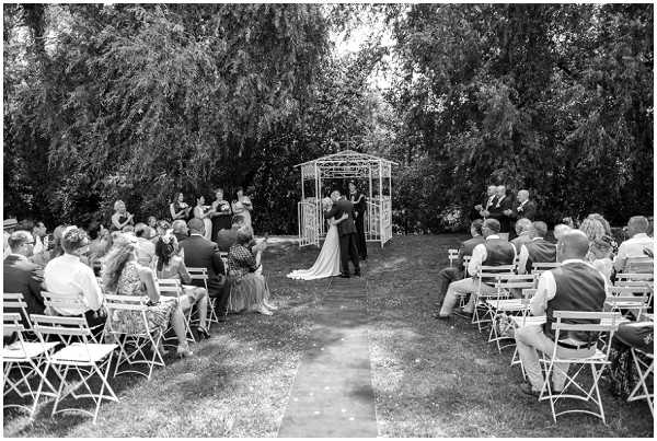 A black-and-white wide shot of an outdoor garden wedding ceremony, capturing the moment the couple shares their first kiss at the altar. The ceremony takes place on a grass lawn with a central aisle runner leading to a white metal gazebo-style arch where the couple and officiant are standing. Approximately 40–50 guests are seated in white folding chairs arranged in two sections on either side of the aisle, with a small bridal party visible to the left of the arch. The contrast is bright and airy, with dark foliage from large trees forming a natural backdrop behind the ceremony space.