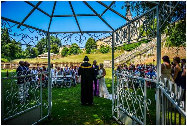 An outdoor wedding ceremony captured from inside a white wrought-iron gazebo, looking out toward the couple and officiant standing before a seated crowd of approximately 40-50 guests arranged in white chairs on a garden lawn. The bride wears a white gown and holds a bouquet, while the officiant wears a black robe with a yellow and black stole. The setting includes a formal garden with stone staircases and balustrades leading up to a historic building with timber-framed architecture visible in the background. A bridesmaid in a pink dress stands to the right side of the frame. The wide-angle shot uses the ornate white ironwork of the gazebo as a natural frame around the ceremony scene.