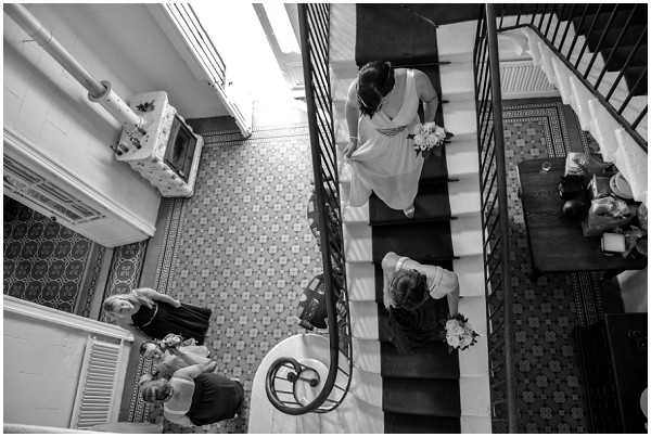Black-and-white overhead shot looking straight down onto an interior staircase of what appears to be a classic French building, featuring ornate patterned floor tiles and a wrought-iron spiral newel post. A bride in a long white gown carries a white bouquet and descends the stairs accompanied by at least one other woman also holding a bouquet, while two more women in dark dresses stand on the tiled landing below. The high-contrast image captures the geometric interplay of the staircase, decorative tilework, and figures from a dramatic bird's-eye perspective. The setting suggests a mairie or historic townhouse interior.