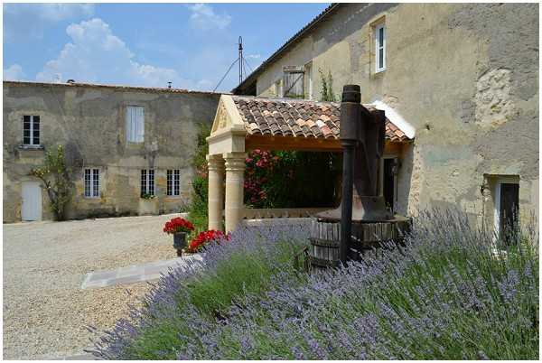 Wide shot of a French country estate courtyard featuring aged stone buildings with white shuttered windows and terracotta roof tiles. A covered wooden wine or olive press structure with a large wooden barrel mechanism is positioned centrally in the courtyard, suggesting a working or historic domaine. Purple lavender plants fill the foreground, and potted red geraniums are placed near the entrance portico with decorative stone columns. No people are present in this image. Potential venue feature image.