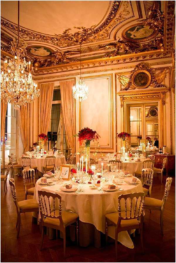 Indoor wedding reception setup in a grand Haussmann-era or palace-style ballroom featuring heavily gilded ceiling moldings, ornate painted medallions, large gold-framed mirrors, and crystal chandeliers casting warm amber light throughout the room. Round tables are dressed in ivory floor-length linens and set with gold Napoléon III-style chairs with tan upholstered seats. Centerpieces consist of tall and low arrangements of deep red flowers, likely red dahlias or red roses, paired with white taper candles in varying heights. Place settings include crystal glassware, white napkins, and small table number frames. The decor palette is rich red, ivory, and gold, giving the room a classic formal style. Wide-angle shot taken from a low perspective, showing multiple tables arranged across the reception space. Potential venue feature image.