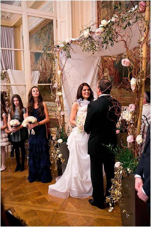 An indoor wedding ceremony taking place in a grand room with ornate painted murals on the walls and a parquet wood floor, consistent with a French chateau or historic mansion. The bride, wearing a white sleeveless high-neck gown, faces the groom who is dressed in a black tuxedo, and she holds a cascading bouquet of ivory and blush flowers. They stand beneath a tall ceremony arch constructed from twisted branches and tall wooden posts, decorated with blush and ivory roses, trailing greenery, and hanging curly willow branches. Two bridesmaids stand to the left — one in a navy tiered ruffle gown and one in a navy sequined dress — with a small guest in a silver sequined top also visible; a bouquet of ivory and cream roses is held by one of the bridesmaids. The ceremony decor features a romantic, organic style with the warm-toned branch structure, soft pink and white floral accents, and a white fabric drape as a backdrop. The shot is a medium wide portrait-style image capturing the couple and the immediate bridal party during the exchange of vows.