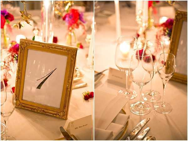 Close-up detail shots of a wedding reception table setting, shown as a diptych. The left image features a gold ornate picture frame displaying the table number '1' in black calligraphy script, surrounded by vibrant fuchsia, red, and magenta floral arrangements, white taper candles in gold holders, and place cards on a white linen tablecloth. The right image shows the same table from a different angle, highlighting multiple crystal wine glasses and a champagne flute, with a lit votive candle, folded white napkins with silverware, and a gold-framed element visible in the background. The decor palette combines gold accents with bold jewel-toned florals, suggesting a formal indoor reception with a classic yet vivid color scheme.