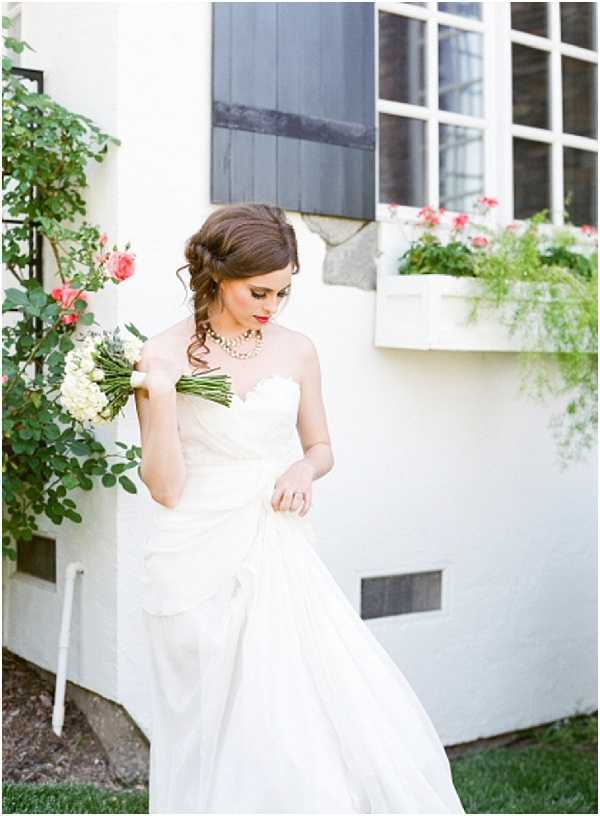 A bridal portrait shot outdoors beside a white-rendered building with dark shuttered windows and a window box filled with coral-pink flowers. The bride wears a strapless ivory ballgown with a sweetheart neckline and full skirt, accessorized with a multi-strand pearl necklace and bold red lipstick, with her dark brown hair styled in a loose updo with soft tendrils. She holds a compact bouquet of cream white flowers, likely white hydrangeas or ranunculus, with tightly wrapped green stems, and looks downward while lifting the hem of her skirt. The styling has a classic, polished feel, and the image is shot on film with warm, soft tones typical of analog photography.