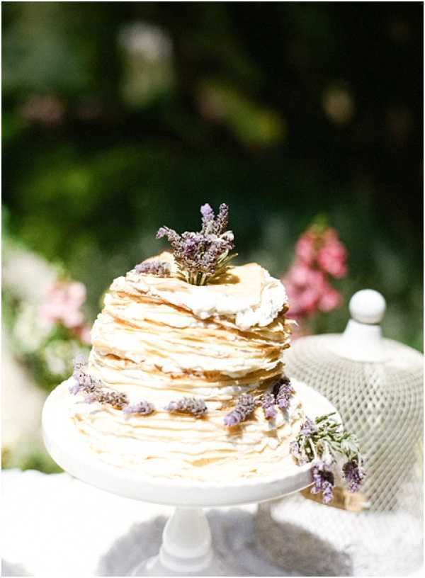 Close-up detail shot of a French crêpe wedding cake stacked in layers with cream filling visible between each crêpe, displayed on a white pedestal cake stand. The cake is decorated with small sprigs of fresh purple lavender placed around the sides and on top, giving it a Provençal rustic style. A white ceramic sugar bowl with a lattice pattern is partially visible to the right, and soft pink florals are blurred in the background, suggesting an outdoor garden setting. The overall decor palette is cream, white, and lavender purple.