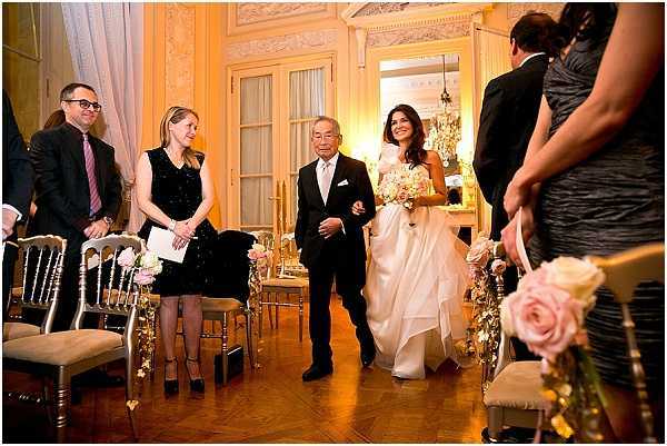 A bride is being walked down the aisle by an older gentleman, presumed to be her father, during an indoor ceremony in what appears to be a ornate Parisian-style salon or chateau ballroom. The bride wears a white ball gown with a full skirt and carries a bouquet of blush pink and ivory flowers. The aisle is lined with gold Chiavari chairs decorated with small floral arrangements featuring blush pink roses and fairy lights. Guests standing along the aisle include a woman in a short black dress and a man in a dark suit with a purple tie, while a bridesmaid in a black dress is visible in the foreground holding a blush pink rose bouquet. The room features ornate molded walls, tall French doors, warm amber lighting from chandeliers, and a parquet wood floor, giving the setting a classic, formal atmosphere. Wide mid-shot taken from a low angle looking up the aisle.