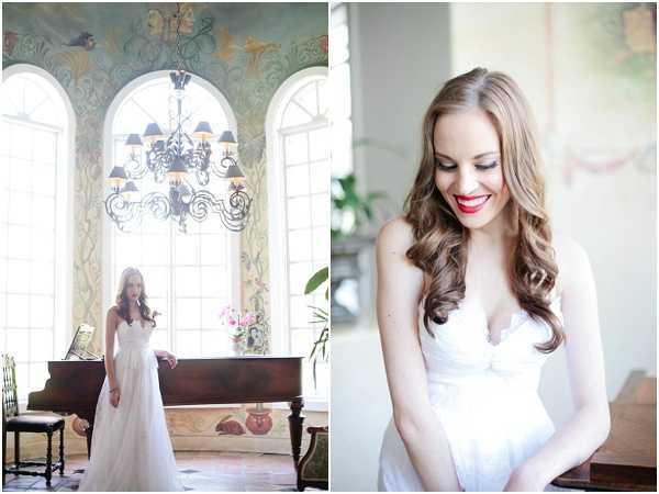 A two-image bridal portrait set taken indoors in an ornate room featuring hand-painted wall murals with botanical and bird motifs, tall arched windows, and a wrought-iron chandelier with black shades. The left image is a wide shot of the bride standing beside a dark wood grand piano, wearing a white strapless chiffon gown with a sweetheart neckline; a small arrangement of pink flowers sits on the piano behind her. The right image is a close-up portrait of the same bride, showing her long side-swept wavy brown hair, bold red lip, and the lace bodice detail of her white gown as she looks downward. Both images share a soft, bright, naturally lit aesthetic consistent with a classic French interior setting.