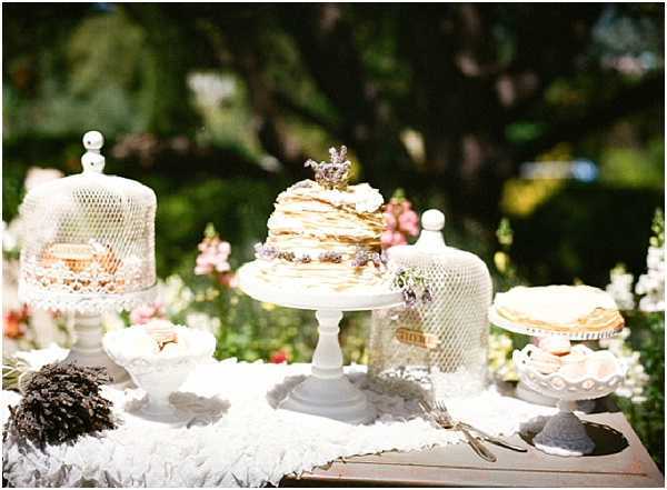 A close-up shot of an outdoor dessert table styled in a vintage, whimsical theme. The centerpiece is a naked crepe cake on a white pedestal stand, topped with small dried lavender sprigs and a vintage figurine topper. Flanking the cake are two white milk-glass dome cloches on matching pedestal stands, covering additional pastries or small cakes. The table is draped in white lace fabric, and vintage silverware is visible in the foreground. A small white milk-glass compote bowl sits to the left. The background is softly blurred greenery and coral-pink flowering plants, suggesting a garden setting. The overall decor palette is ivory and white with muted lavender accents, consistent with a rustic or vintage-inspired wedding aesthetic.