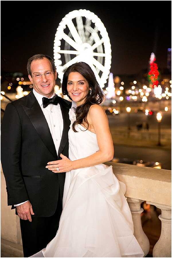 A couple portrait taken outdoors at night on a stone balustrade terrace, with the bride and groom posing together facing the camera. The groom wears a black tuxedo with a black bow tie and white dress shirt, while the bride wears a strapless white ballgown with a voluminous skirt. Behind them, a large illuminated Ferris wheel is prominently lit in white against the dark sky, surrounded by bokeh city lights, placing the setting in central Paris — consistent with the Place de la Concorde area. The composition is a mid-length portrait shot with a shallow depth of field that blurs the city background into warm glowing orbs of light.