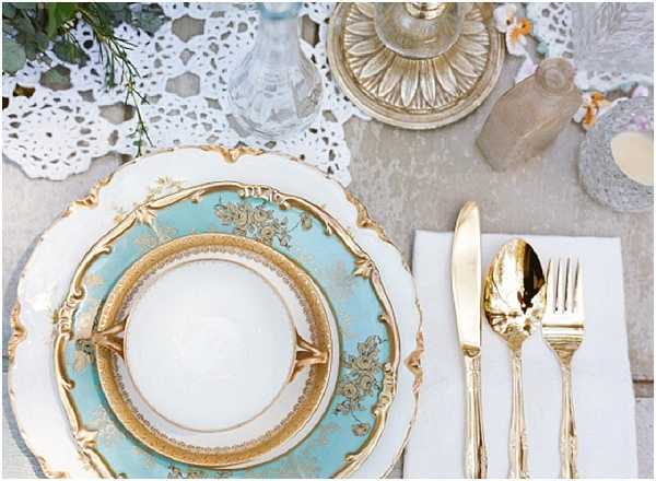 A close-up detail shot of a vintage-style wedding table setting featuring a stacked place setting of white and duck-egg blue china plates with ornate gold floral detailing, layered on a white crocheted lace doily. Gold flatware — knife, fork, and spoon — is laid on a white linen napkin to the right. Background elements include a gold lidded compote dish, small clear glass bud vases, a frosted glass bottle, and a small candle, all arranged on a neutral grey linen tablecloth. The overall decor palette combines soft duck-egg blue, white, and gold, with a vintage or antique-inspired styling theme.