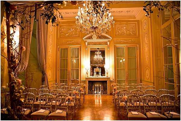 An indoor ceremony space set up in a grand classical French salon, empty and awaiting guests. The room features ornate gold-painted wall paneling with bas-relief decorations, tall French doors with sage green curtains, and a marble fireplace serving as the focal point at the far end. Two large crystal chandeliers hang from the coffered ceiling, casting warm amber light throughout the space. Rows of gold chiavari chairs are arranged on either side of a central aisle, each dressed with small floral arrangements in peach and blush tones, while tall branching arrangements with delicate pink florals flank the aisle entrance. Candelabras with white taper candles are placed along the aisle and on the fireplace mantel, reinforcing a warm candlelit atmosphere with a classic, ornate French interior style. Wide shot capturing the full length of the ceremony setup. Potential venue feature image.
