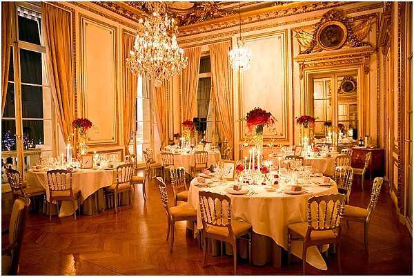 A wedding reception dinner setup inside a grand Haussmann-style Parisian ballroom, photographed at night with city lights visible through tall windows. The room features ornate gold molding, crystal chandeliers, large mirrors with gilded frames, and floor-length gold satin drapes. Round tables are dressed in ivory or cream floor-length linens and set for approximately 8–10 guests each, with crystal glassware, white candles in tall candlesticks, and deep red floral centerpieces — likely red roses. Gold Napoléon III-style chairs surround each table. The warm amber lighting from the chandeliers and candles creates a rich, golden tone throughout the room. Wide-angle interior shot showing the full reception setup with no guests present. Potential venue feature image.