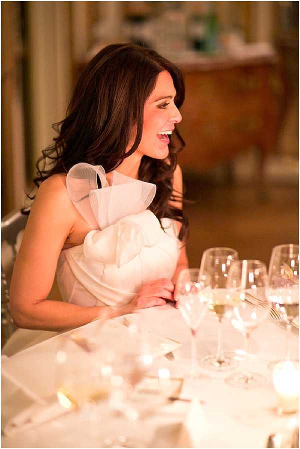 A bride is seated at what appears to be the head table during an indoor wedding reception, laughing and looking to one side. She is wearing a white one-shoulder gown with a large structured organza ruffle detail at the neckline and chest. Her dark brown hair falls in loose waves over her shoulders. The table in front of her is set with white linens, crystal wine glasses, and small votive candles providing warm ambient lighting. The background is softly blurred, revealing ornate gilded furniture consistent with a chateau or formal ballroom setting. This is a mid-range portrait shot with warm candlelit tones.