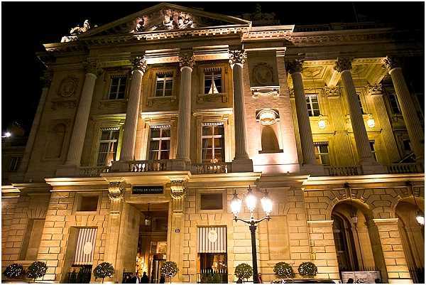 Night exterior shot of a grand neoclassical building, identified by a sign above the entrance as the Hôtel de Crillon, photographed from street level looking up at the illuminated façade. The building features tall Corinthian columns, ornate stonework, a pediment, and multiple floors of tall windows, all lit with warm golden uplighting against a dark night sky. A traditional multi-armed street lamp stands in front of the entrance, and topiary bushes in pots flank the doorway. Wide architectural shot with no people prominently visible. Potential venue feature image.