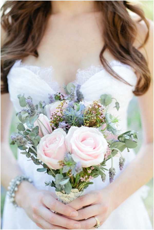 Close-up portrait of a bride holding a small, loosely arranged bouquet featuring two large blush pink roses, blue eryngium thistle, lavender wax flower, eucalyptus, and soft purple filler blooms. The bride wears a white strapless sweetheart-neckline gown with lace or ruffle detailing at the bust, a gold ring on her finger, and a pearl or crystal bracelet on her wrist. The bouquet stems are wrapped in what appears to be lace or hessian ribbon. The overall floral palette of blush, dusty blue, and sage green suggests a soft romantic styling with a garden-gathered, boho aesthetic.