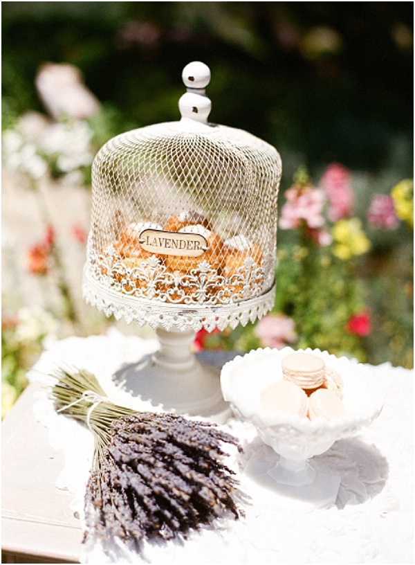 A close-up detail shot of an outdoor wedding dessert or favor table styled with a French Provençal theme. The centerpiece is a white ornate wire mesh cloche on a white pedestal stand, labeled 'Lavender' and containing small baked goods or favors. To the right sits a white milk glass compote dish holding pale pink and ivory macarons. In the foreground, a tied bundle of dried lavender sprigs rests on the white table surface. Blurred pink, red, and yellow florals fill the background, suggesting a garden setting. The overall decor palette is white and soft neutrals with the dried lavender adding muted purple tones, consistent with a rustic French countryside styling theme.