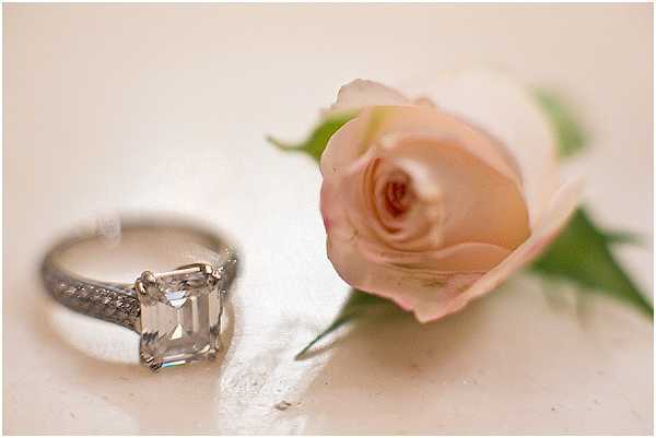 A close-up detail shot of a silver engagement ring featuring a large emerald-cut diamond solitaire with a pavé-set band, placed on a soft white surface beside a single blush pink rose boutonniere with green foliage. The shallow depth of field keeps the ring in sharp focus while the rose is slightly soft in the background. The overall palette is pale and muted, with warm ivory tones in the surface and soft pink in the rose.