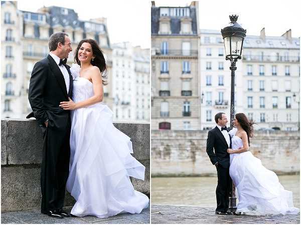 A side-by-side couple portrait session along the Seine River in Paris, with classic Haussmann-style apartment buildings visible in the background. The bride wears a strapless white ballgown with a voluminous, layered skirt that is visibly caught in the wind, paired with drop earrings and dark hair worn down. The groom wears a classic black tuxedo with a bow tie. The left image shows the couple laughing together while leaning against a stone quay wall, and the right image shows them standing close together beside a traditional Parisian cast-iron street lamp on the riverbank. Both shots are outdoor portraits taken at ground level with a shallow depth of field.