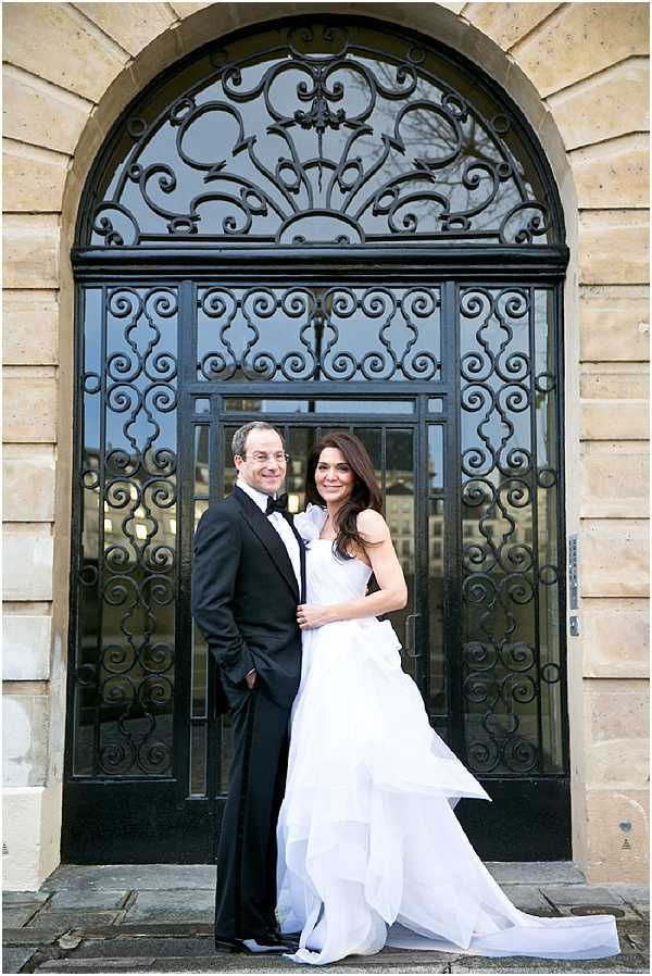 A couple portrait taken outdoors in front of a large ornate arched doorway featuring elaborate black wrought-iron scrollwork set into a pale stone building facade. The bride wears a white ballgown with a ruffled one-shoulder detail and a full, layered skirt with a train, while the groom is dressed in a classic black tuxedo with a bow tie. The two stand close together on stone steps, both facing the camera. The composition is a mid-range portrait shot with the decorative door as a strong architectural backdrop. Potential venue feature image.