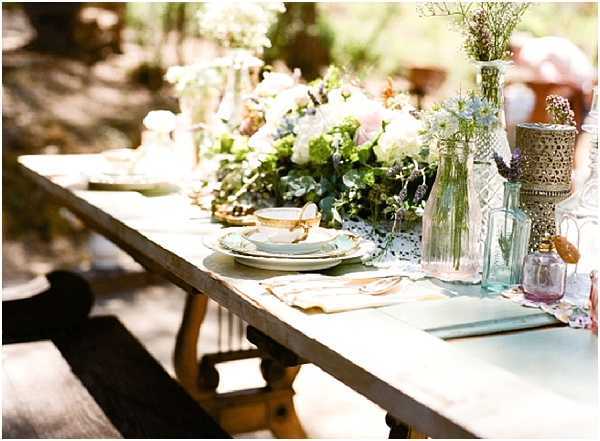 Close-up detail shot of an outdoor reception table set on a rustic wooden picnic-style trestle table, styled in a boho-vintage aesthetic. The place setting features mismatched vintage china with a gold-rimmed teacup and saucer stacked on layered plates, accompanied by a linen napkin in soft yellow tones. A lush floral centerpiece runs along the table featuring white and blush roses, greenery, and eucalyptus, surrounded by an eclectic mix of clear glass bottles, a pale blue vintage bottle, a lavender glass vessel, and an ornate gold lantern or vessel, all arranged loosely along a pale mint runner. The setting is outdoors in a dappled, shaded garden environment, and the overall decor palette combines mint, blush, gold, and soft lavender in a whimsical, mismatched vintage style.