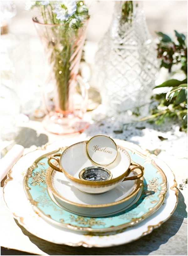 Close-up detail shot of a vintage-style wedding table setting featuring a stacked place setting with a pale robin's egg blue and gold-rimmed decorative charger plate topped with a white dinner plate, a gold-rimmed teacup and saucer, and a small round place card holder with script calligraphy reading 'Karolina.' A compass or small decorative object sits inside the teacup. In the soft-focus background, a blush pink pressed glass vase holds greenery and small blue flowers, alongside a cut crystal decanter or vase. The overall decor palette combines robin's egg blue, gold, blush pink, and white in a vintage, antique-inspired styling theme. The table linen appears to be white with a lace or textured overlay.