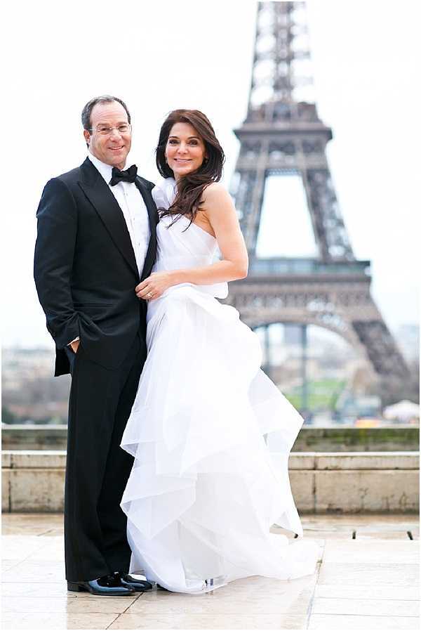A couple poses for a portrait outdoors at the Trocadéro in Paris, with the Eiffel Tower prominently centered in the background. The bride wears a strapless white ballgown with layered, ruffled organza skirt panels, and her dark hair is worn loose and flowing; the groom is dressed in a classic black tuxedo with a black bow tie and patent leather shoes. The composition is a mid-length portrait shot with the couple standing close together, both facing the camera and smiling. The styling is formal and classic, with an overcast white sky providing soft, diffused natural light.