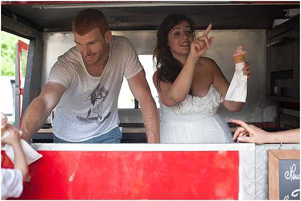 A candid reception moment showing the bride and a male guest — likely the groom in casual attire — serving from inside a red food truck or ice cream van. The bride wears a strapless white dress with ruffled bodice detail and holds an ice cream cone while pointing upward, laughing. The man wears a white graphic t-shirt and jeans. Guests' hands are visible reaching up to the service window on the right. A small chalkboard menu sign is partially visible in the lower right corner. Medium shot taken from outside the truck at window level, with a relaxed, informal atmosphere suggesting a casual outdoor cocktail or reception catering moment.