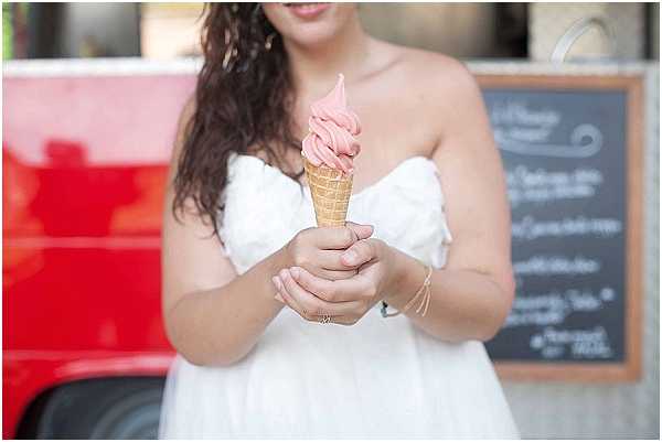 A woman in a white off-the-shoulder dress holds a pink soft-serve ice cream cone in a waffle cone with both hands at a cocktail hour or reception activity. She is standing in front of what appears to be a red food truck or ice cream van, with a chalkboard menu board visible in the background. The shot is a mid-range portrait cropped at the chest, with focus on the ice cream cone and her hands, which feature delicate gold bracelets. The styling is casual and relaxed, suggesting a fun catering element incorporated into the wedding event.